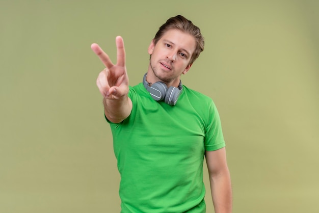 Young handsome man wearing green t-shirt with headphones smiling showing and pointing with fingers number two or victory sign standing over green wall