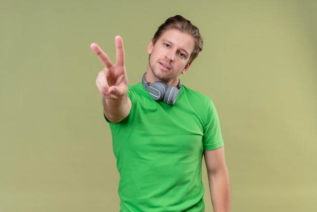 Молодой красавец в зеленой футболке с наушниками улыбается, показывает пальцами номер два или показывает знак победы, стоящий над зеленой стеной