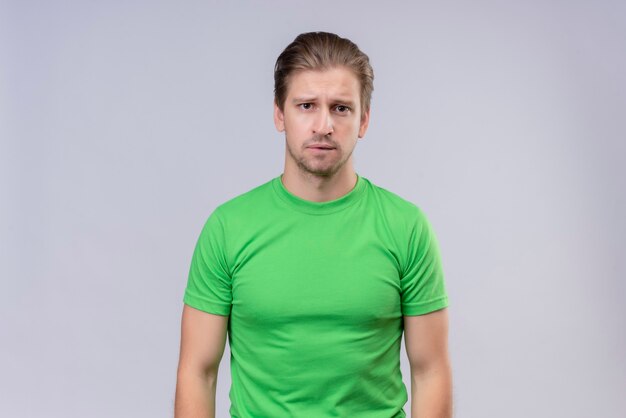 白い壁の上に立っている顔に悲しそうな表情で緑のtシャツを着ている若いハンサムな男