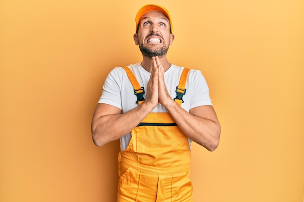 Young handsome man wearing handyman uniform over yellow background begging and praying with hands together with hope expression on face very emotional and worried begging