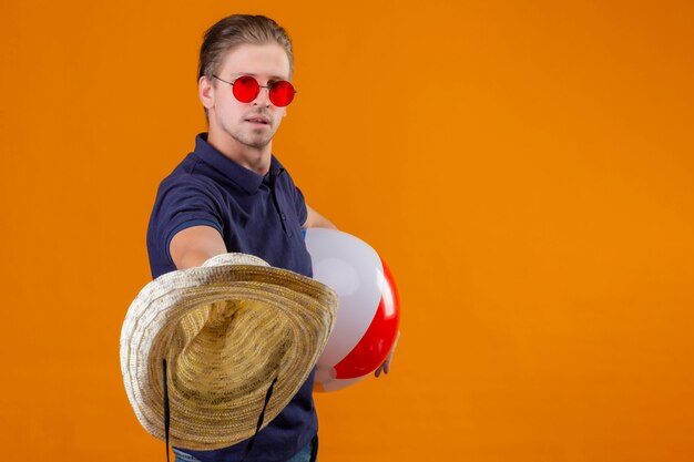 膨らませてボールを押しながらオレンジ色の背景の上に立っている自信を持って式で麦わら帽子を伸ばして赤いサングラスをかけている若いハンサムな男