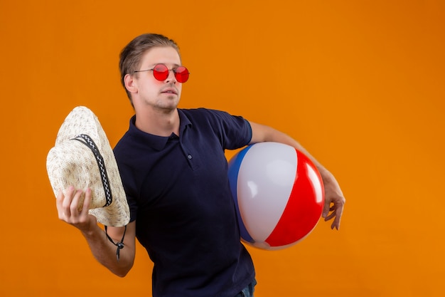 Free photo young handsome man wearing red sunglasses standing with inflatable ball waving with straw hat looking confident over orange background