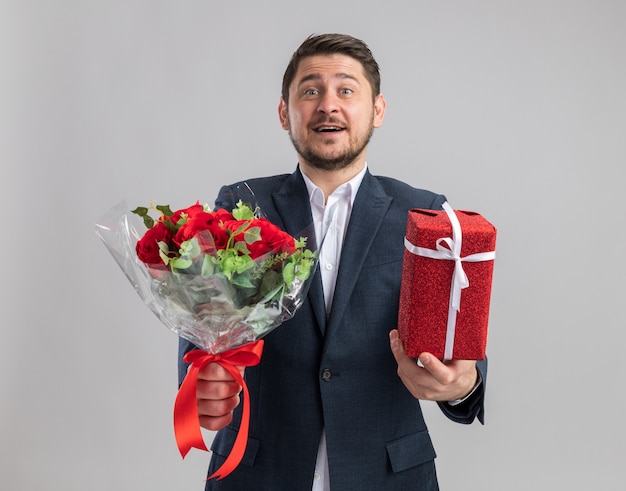 Giovane uomo bello che indossa un abito con un mazzo di rose e un regalo per san valentino felice ed eccitato in piedi sul muro bianco white