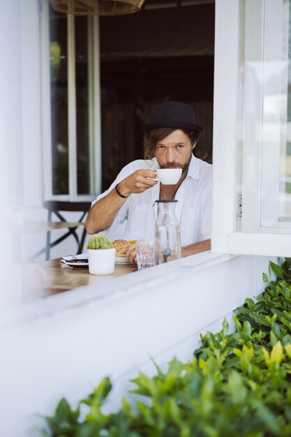 Young handsome man in a white shirt open, having breakfast in a cafe with a vegetarian burger, drinking coffee, lifestyle in a tropical island, life in Bali.