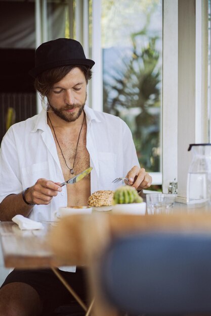 白いシャツを着た若いハンサムな男が開いて、菜食主義のハンバーガーと一緒にカフェで朝食をとり、コーヒーを飲み、熱帯の島でのライフスタイル、バリでの生活。