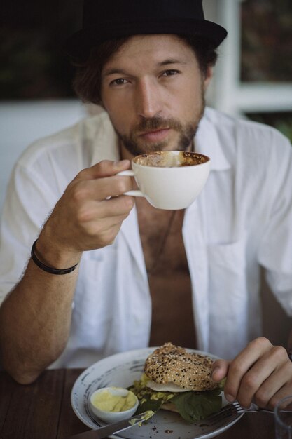 Молодой красивый мужчина в открытой белой рубашке, завтракает в кафе с вегетарианским бургером, пьет кофе, образ жизни на тропическом острове, жизнь на Бали.