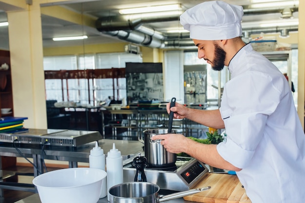 パスタを準備している白い特別なローブを着た若いハンサムな男は、イタリアのスパゲッティのために水を沸騰させます。