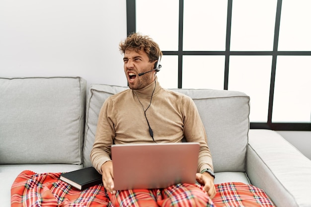 Young handsome man with beard wearing operator headset working from home angry and mad screaming frustrated and furious shouting with anger rage and aggressive concept