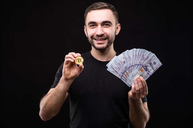 Young handsome man with golden bitcoin and with dollar cash in other hand isolated on black