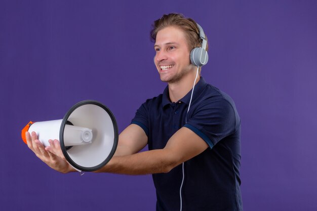 Young handsome man with headphones holding megaphone with big smile on face standing over purple background