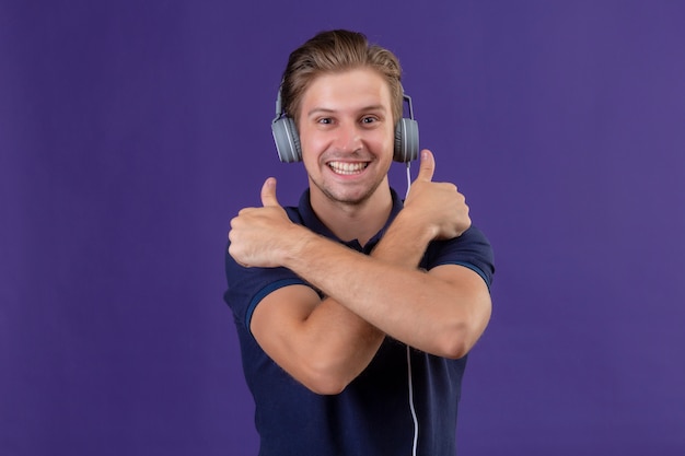 Young handsome man with headphones standing with arms crossed showing thumbs up smiling cheerfully over purple background