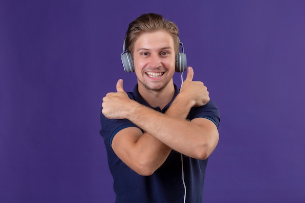 腕を組んで立っているヘッドフォンで若いハンサムな男交差紫色の背景に元気に笑って親指を表示