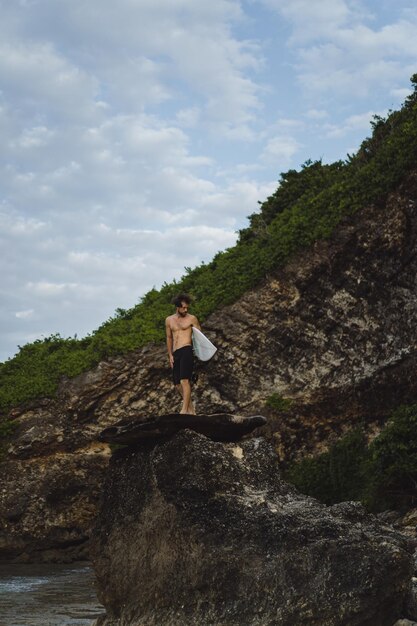 海の近くの岩の上にサーフボードを持つ若いハンサムな男。