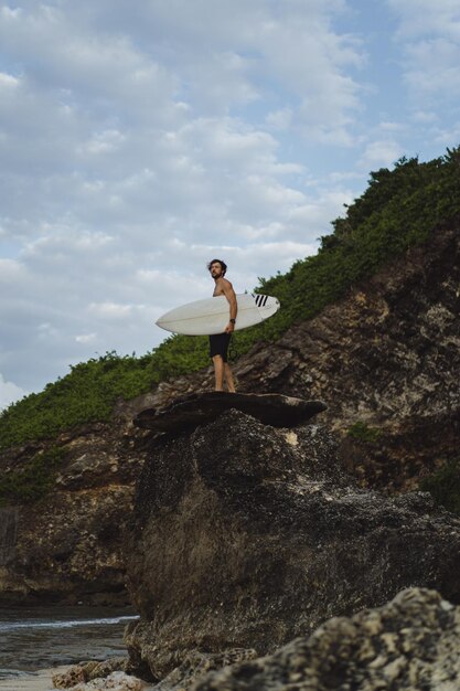 海の近くの岩の上にサーフボードを持つ若いハンサムな男。