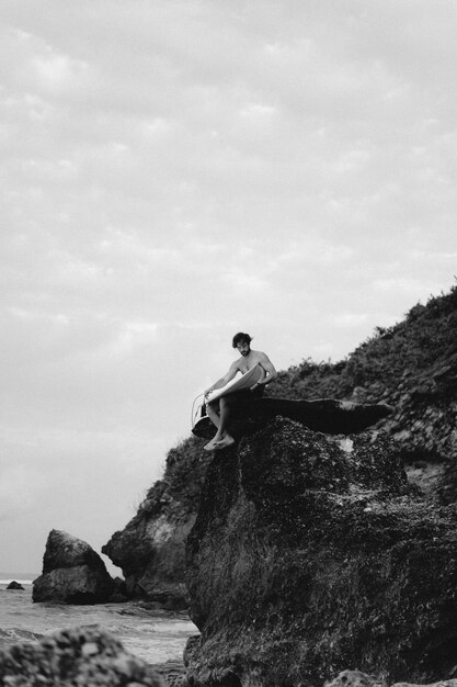 Young handsome man with a surfboard on a rock near the ocean.