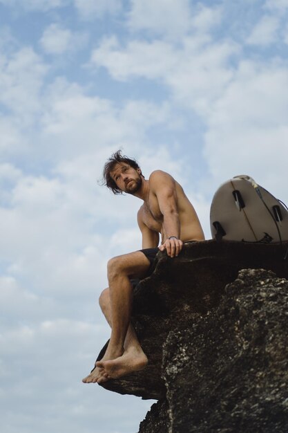 Young handsome man with a surfboard on a rock near the ocean.