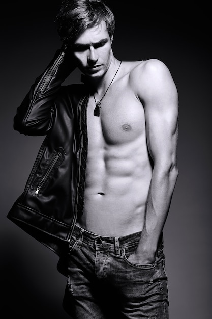 Young handsome muscled fit male model man in leather jacket posing in studio showing his abdominal muscles