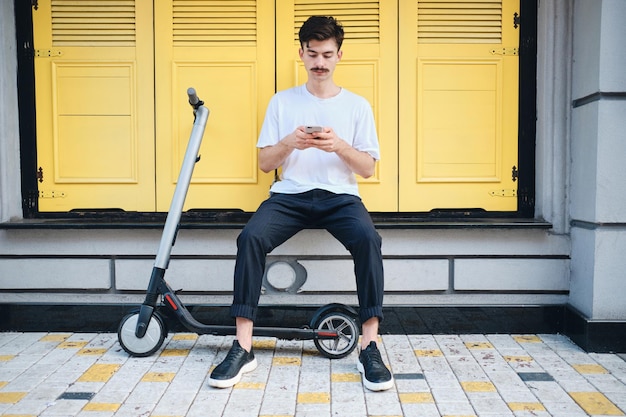 Young handsome mustache man sitting with e-scooter while thoughtfully using cellphone with yellow background on city street