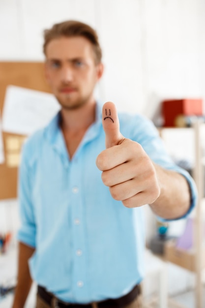 Free photo young handsome pensive businessman showing thumb up with funny sad face drawing focus on hand white modern office interior background