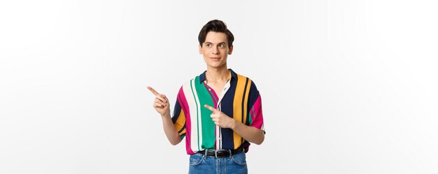 Young handsome queer man pointing looking left at logo smiling pleased standing over white backgroun