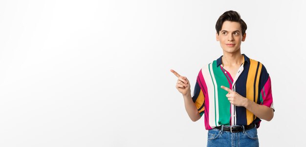 Young handsome queer man pointing looking left at logo smiling pleased standing over white background