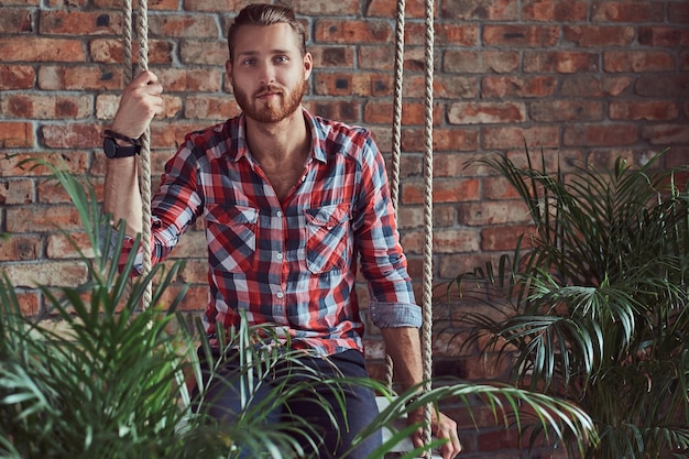 Free photo a young handsome redhead model man sits on a swing in the room with a loft interior.