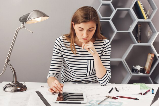 Young handsome serious engineer girl with long brown hair in striped shirt holding head with hand, looking in papers with serious face expression, trying to find a mistake in blueprints.