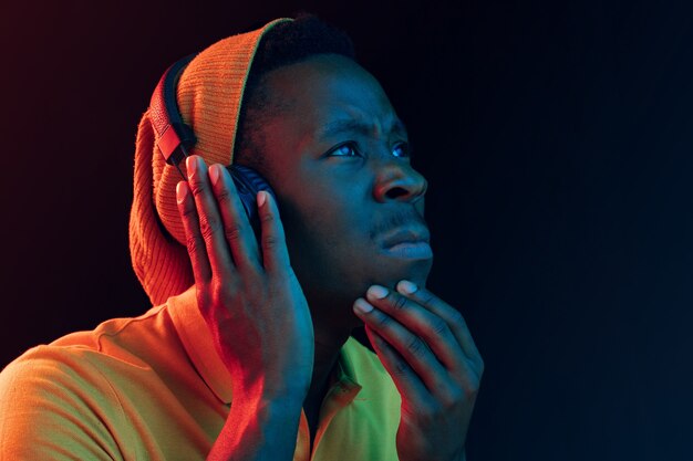 The young handsome serious sad hipster man listening music with headphones at black studio with neon lights. Disco, night club, hip hop style, positive emotions, face expression, dancing concept