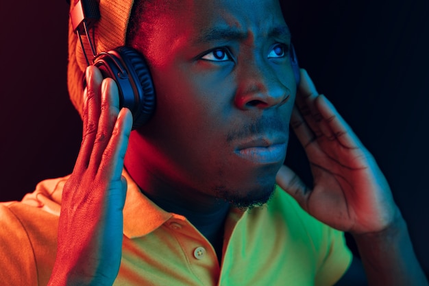 The young handsome serious sad hipster man listening music with headphones at black studio with neon lights. Disco, night club, hip hop style, positive emotions, face expression, dancing concept