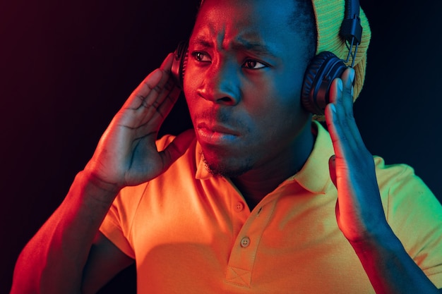 Молодой красивый серьезный грустный хипстерский мужчина слушает музыку в наушниках в черной студии с неоновыми огнями
