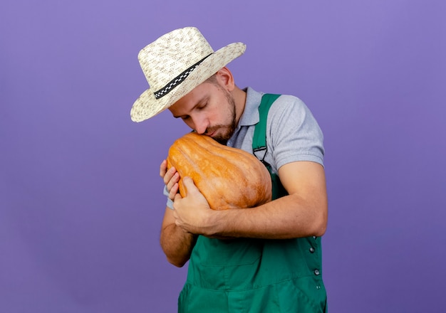 제복을 입은 젊은 잘 생긴 슬라브 정원사 및 복사 공간이 보라색 벽에 고립 된 닫힌 눈을 가진 butternut 호박을 들고 모자