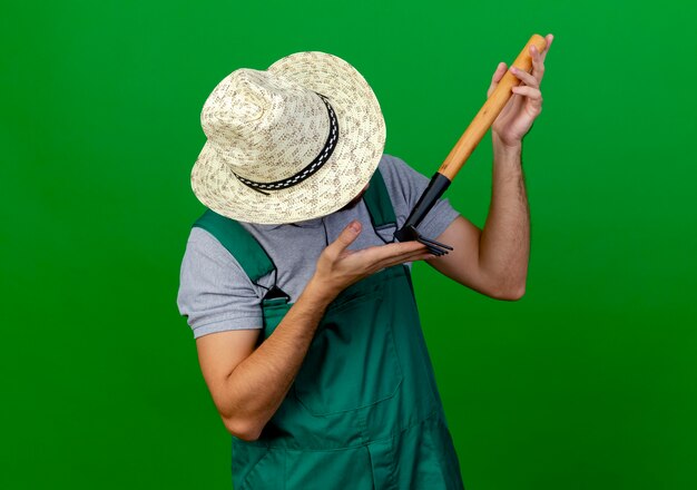 Young handsome slavic gardener in uniform and hat holding and looking at rake 