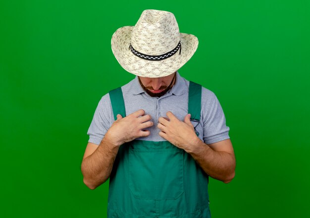 Young handsome slavic gardener in uniform and hat touching chest and looking down at it 