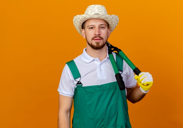 Young handsome slavic gardener in uniform wearing hat and gardening gloves holding pruners on shoulder looking  isolated on orange wall with copy space