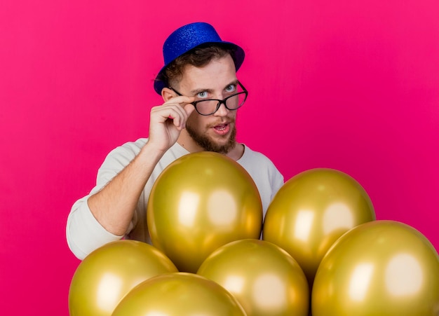 Ragazzo di partito slavo bello che indossa cappello e occhiali da festa in piedi dietro palloncini afferrando occhiali guardando davanti isolato sul muro rosa