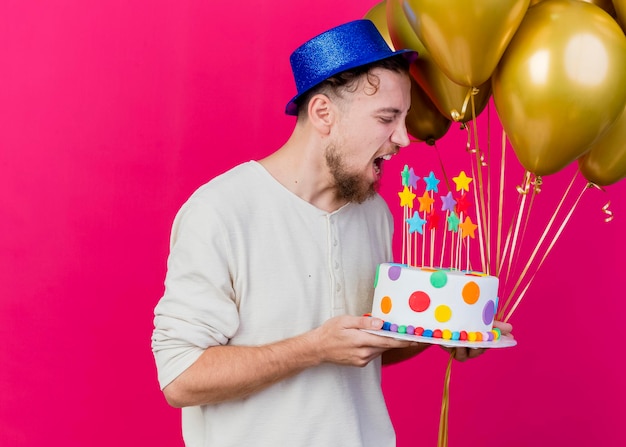 Молодой красивый славянский тусовщик в шляпе с воздушными шарами и праздничным тортом со звездами готовится откусить торт, изолированный на розовой стене с копией пространства