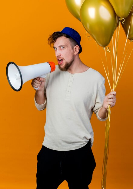 Young handsome slavic party guy wearing party hat holding balloons looking at camera talking by speaker isolated on orange background