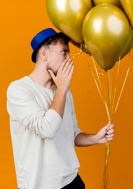 Young handsome slavic party guy wearing party hat holding balloons looking straight whispering isolated on orange wall