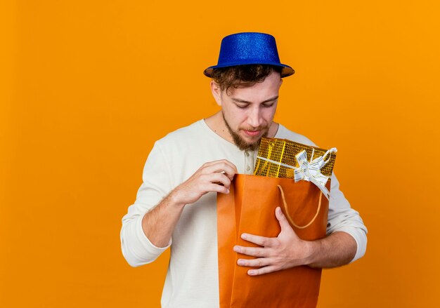 Young handsome slavic party guy wearing party hat holding and looking inside paper bag with gift boxes isolated on orange background with copy space