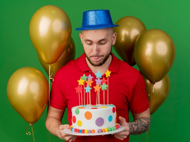 Free photo young handsome slavic party guy wearing party hat standing in front of balloons holding and looking at birthday cake and showing tongue isolated on green background