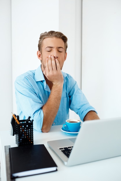 Young handsome sleepy confident pensive businessman sitting at table working on laptop with cup of coffee aside. White modern office interior 