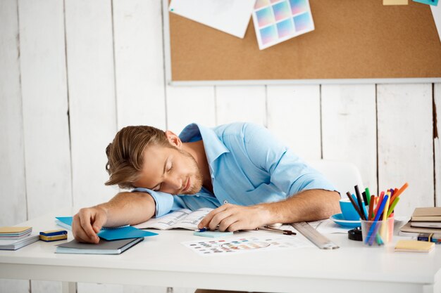 Young handsome sleepy tired businessman sleeping at table on papers and notepad. White modern office interior 