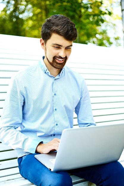 Young handsome smiling businessman model sitting on the park bench using laptop in casual hipster cloth