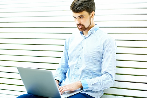 Young handsome smiling businessman model sitting on the park bench using laptop in casual hipster cloth