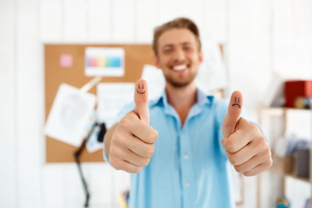 Young handsome smiling businessman showing thumbs up with funny faces drawings. Focus on hands.