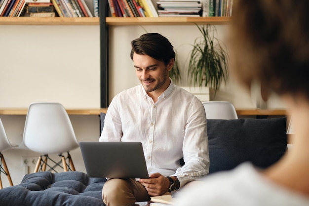 Young handsome smiling businessman sitting on sofa with colleague while happily working on laptop in modern coworking space