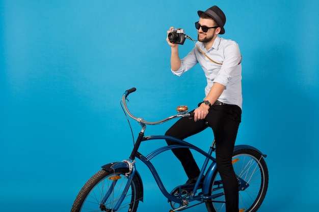 若いハンサムな笑顔幸せな男の流行に敏感な自転車で旅行、青いスタジオの背景にビンテージ写真のカメラを保持している、シャツ、帽子、サングラス、写真家が写真を撮る