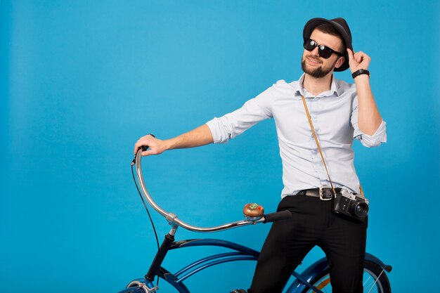 Young handsome smiling happy man traveling on hipster bicycle, posing on blue studio background, wearing shirt, hat and sunglasses