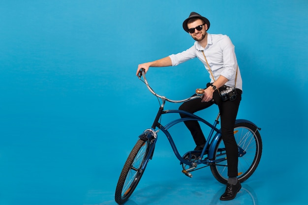 Young handsome smiling happy man traveling on hipster bicycle, posing on blue studio background, wearing shirt, hat and sunglasses