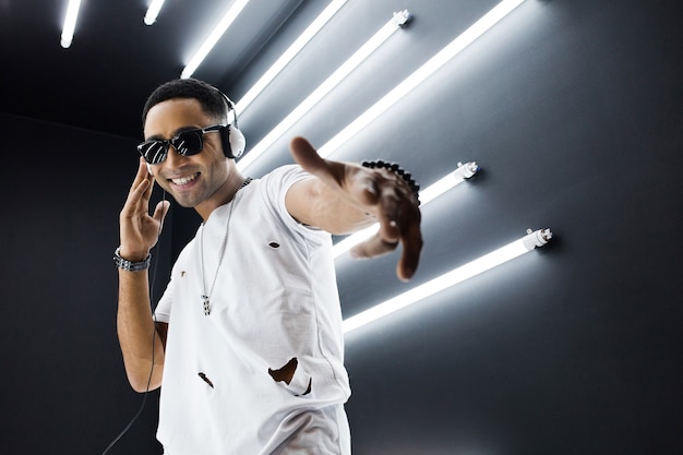 Young handsome smiling hipster black man in white outfit listening to music on headphones and dancing in hip hop style in disco night club, having fun